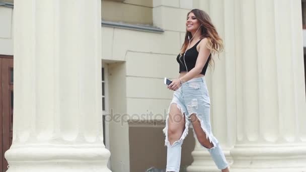 Девушка в разорванных джинсах и футболках, гуляющая по летнему городу и слушающая музыку в наушниках. замедленное движение — стоковое видео