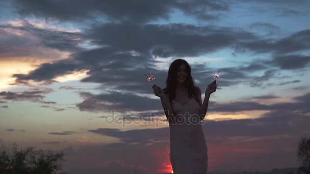 Mooi sexy meisje in vecherom jurk dansen op het strand bij zonsondergang. een jonge vrouw houdt de wonderkaarsen en plezier hebben op de partij. Slow motion — Stockvideo