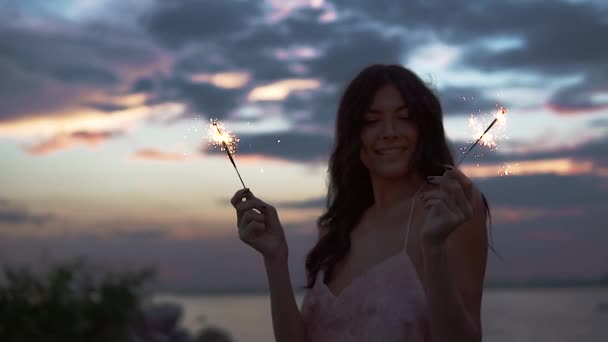 Portret van een verleidelijke meisje met wonderkaarsen bij zonsondergang. Mooie jonge vrouw dansen op de kust. Slow motion — Stockvideo