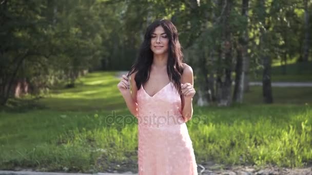 Porträt einer Brünetten in einem Cocktailkleid vor einem Hintergrund grüner Bäume. junges Mädchen posiert und lächelt in die Kamera — Stockvideo