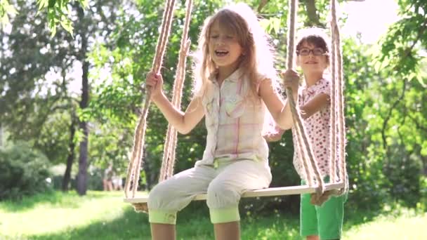 Niños balanceándose en un columpio en el parque de verano. dos hermanitas jugando al aire libre. la hermana mayor sacude a su hermana menor en el columpio. columpio en las cuerdas unidas al árbol — Vídeo de stock
