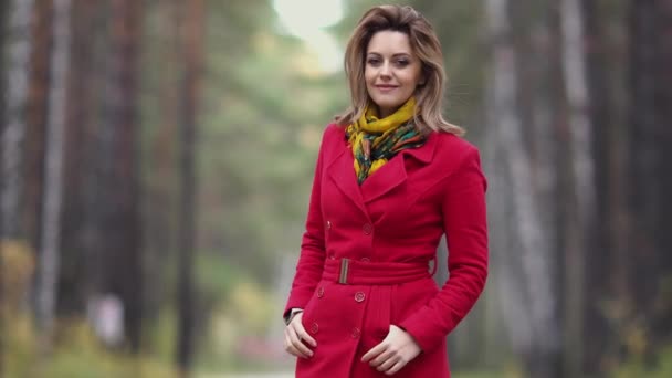 Close-up portret van een mooie jonge vrouw op een achtergrond van herfst bos. meisje in de rode jas glimlacht en kijkt naar de camera — Stockvideo