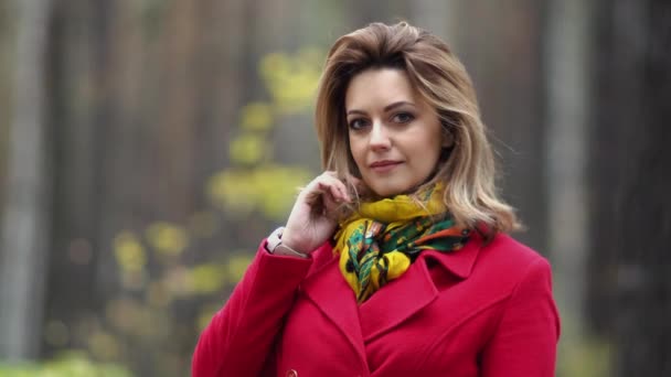 Close-up portret van een mooie jonge vrouw op een achtergrond van herfst bos. meisje in de rode jas glimlacht en kijkt naar de camera — Stockvideo