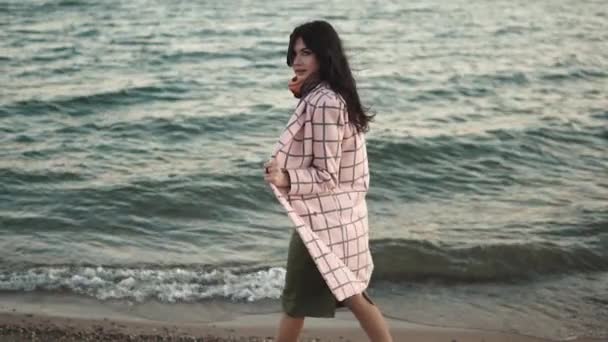 Linda chica en abrigo de otoño camina a lo largo de la playa al atardecer. chica posando para la cámara al aire libre. Movimiento lento — Vídeos de Stock