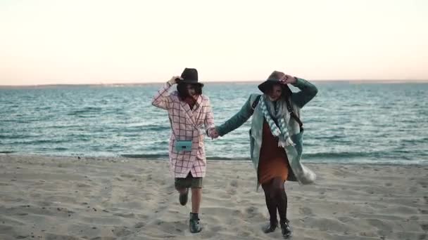 Freundinnen laufen fröhlich am Strand entlang und werfen ihre Hüte hoch. schöne junge Frauen genießen einen herbstlichen Nachmittag im Freien. Zeitlupe — Stockvideo