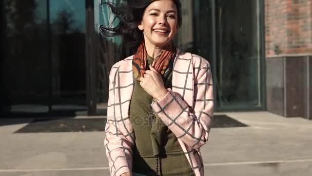 Charmante jongedame doorloopt de herfst stad in een jas en glimlacht. leuk meisje met een boeket bloemen op een achtergrond van moderne architectuur. Slow motion — Stockvideo