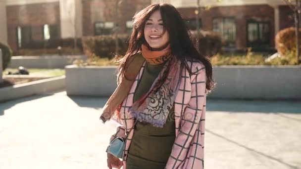 Uroczy młoda kobieta idzie przez miasto jesień w płaszcz i uśmiecha się. Ładna dziewczyna na tle współczesnej architektury. zwolnionym tempie — Wideo stockowe