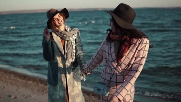 Beztroski dziewczyna chodzić po plaży o zachodzie słońca i cieszyć się ciepły, jesienny wieczór. dwie dziewczyny w płaszcz jesień bawimy się razem. zwolnionym tempie — Wideo stockowe
