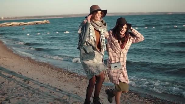 Dos novias corren alegremente y se divierten al aire libre. mujer joven en abrigo de otoño y sombrero en la playa al atardecer. Movimiento lento — Vídeos de Stock