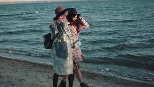 Novia despreocupada caminar por la playa al atardecer y disfrutar de la cálida noche de otoño. dos chicas en abrigo de otoño se divierten juntas. cámara lenta — Vídeos de Stock