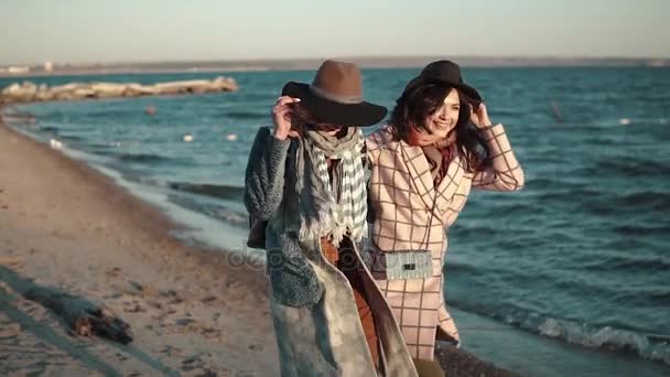 Dos novias corren alegremente y se divierten al aire libre. mujer joven en abrigo de otoño y sombrero en la playa al atardecer. Movimiento lento — Vídeos de Stock