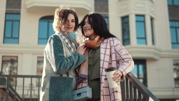 Retrato de dos mujeres jóvenes lindas en abrigo de otoño sobre un fondo de la arquitectura moderna. mejores amigos juntos — Vídeo de stock
