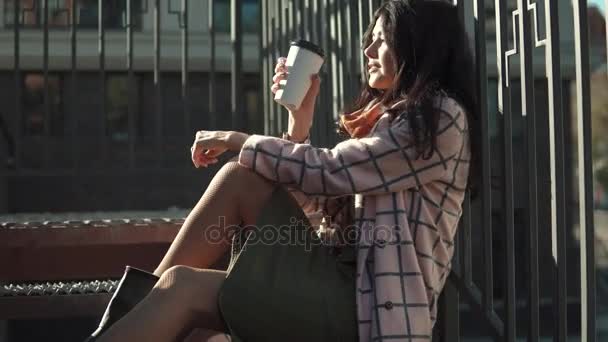 Портрет милой молодой женщины на фоне осеннего города. Девушка в осеннем пальто пьет кофе из бумажной чашки и наслаждается теплым осенним днем — стоковое видео