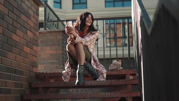 Retrato de una linda joven sobre el telón de fondo de una ciudad otoñal. una chica en un abrigo de otoño bebe café de una taza de papel y disfruta de un cálido día de otoño — Vídeos de Stock