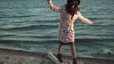 sonbahar paltolu sevimli kız batımında sahilde yürüyor. genç kadının su üzerinde sıcak sonbahar günü. Kız komik dengesini korumak için bir oturum açma gider