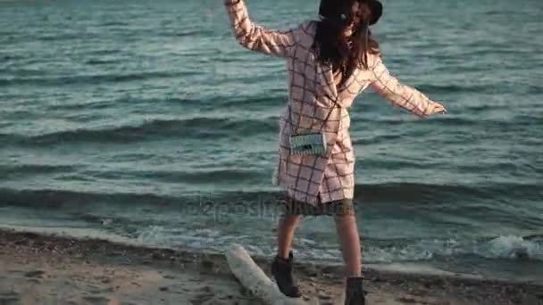 가 코트에 귀여운 여자 해질녘 해변을 따라 산책. 젊은 여자는 물에 따뜻한가 날을 즐기고. 여자 재미는 균형을 유지 하려고 하는 로그에 간다 — 비디오
