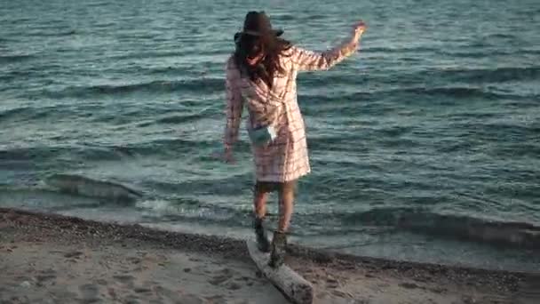 Nettes Mädchen im Herbstmantel spaziert bei Sonnenuntergang am Strand entlang. junge Frau genießt warmen Herbsttag auf dem Wasser. das Mädchen geht lustig auf einem Baumstamm herum und versucht das Gleichgewicht zu halten — Stockvideo