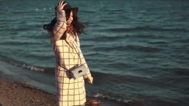 Jonge vrouw genieten van een warme herfst middag door het water. meisje in herfst jas en hoed wandelen langs de kust bij zonsondergang — Stockvideo