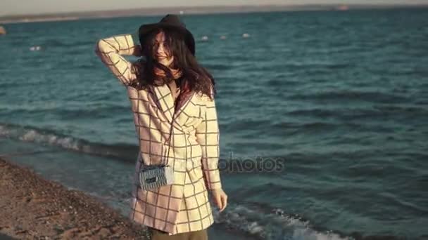 Молодая женщина наслаждается теплым осенним днем у воды. девушка в осеннем пальто и шляпе, идущая вдоль побережья на закате — стоковое видео