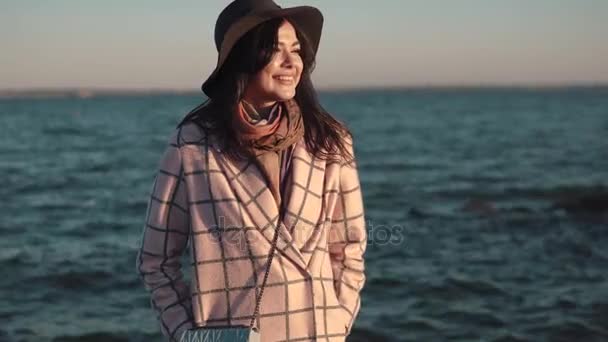 Portrait of a young woman on the beach. girl in hat and autumn coat smiling and posing on camera by the water. — Stock Video