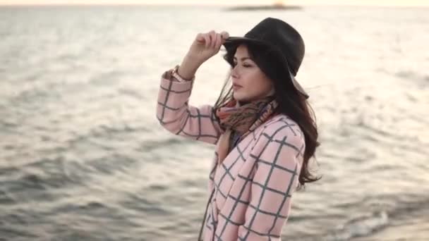 Porträtt av en ung kvinna på stranden. flicka i hatt och höst kappa leende och poserar på kameran vid vattnet. — Stockvideo