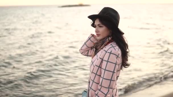 Ritratto di una giovane donna sulla spiaggia. ragazza in cappello e cappotto autunnale sorridente e posa sulla macchina fotografica vicino all'acqua . — Video Stock