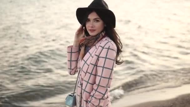 Sahilde genç bir kadın portresi. Kız gülümseyerek ve su kamera poz şapka ve sonbahar ceket. — Stok video