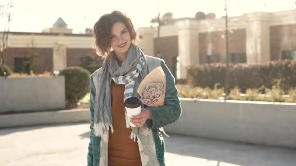 Jonge vrouw met een boeket bloemen en een kopje koffie poseren op de camera op de achtergrond van stedelijke architectuur. modieuze meisje in een herfst jas — Stockvideo