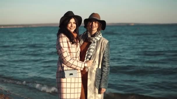 Dos mujeres jóvenes lindas sonríen y disfrutan de la vida en un paseo de otoño a lo largo del océano. retrato de amigos en el fondo del mar de otoño — Vídeos de Stock