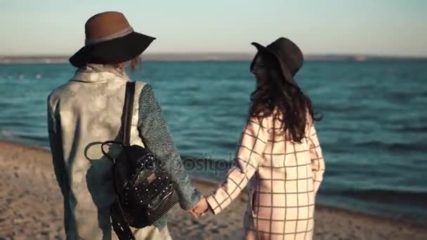 Migliori amici si tengono per mano e camminare lungo la spiaggia al tramonto. le ragazze stanno godendo di una calda giornata autunnale — Video Stock