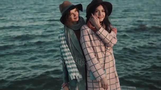 Portrait of charming girl in hat and coat at sunset on the background of the autumn sea. friends spend time outdoors — Stock Video