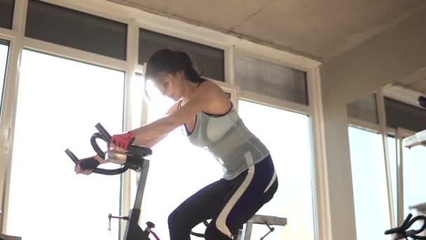Vonzó nő fokozása her tartóssági, miközben dolgozik egy exercycle. egy gyönyörű lány, az edzőteremben, helyhez kötött kerékpár portré. fiatal nő a gyakorlás kerékpár aerob testmozgás — Stock videók