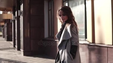 sevimli genç kadın bir ceket ve gülümsüyor sonbahar şehirde size yürür. modern mimarinin bir arka plan üzerinde şirin kız. ağır çekim