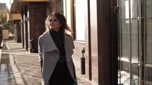 Affascinante giovane donna cammina attraverso la città autunnale in un cappotto e sorride. ragazza carina su uno sfondo di architettura moderna. rallentatore — Video Stock