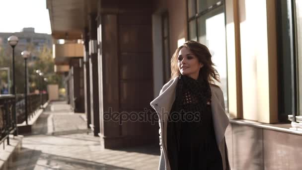 Чарівна молода жінка гуляє по осінньому місту в пальто і посміхається. мила дівчина на тлі сучасної архітектури. повільний рух — стокове відео