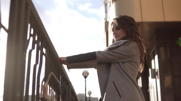 Sevimli genç kadın bir ceket ve gülümsüyor sonbahar şehirde size yürür. modern mimarinin bir arka plan üzerinde şirin kız. ağır çekim — Stok video