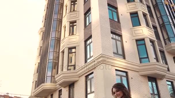 Charmante jeune femme marche à travers la ville d'automne dans un manteau et sourit. fille mignonne sur un fond d'architecture moderne . — Video