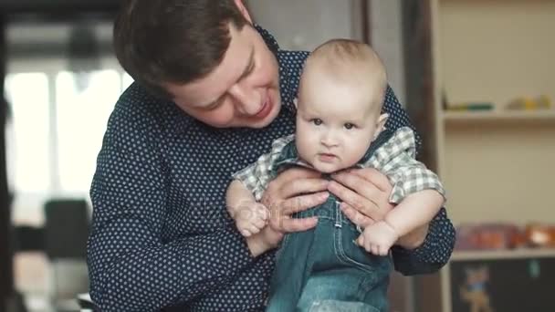 Ung pappa håller sin unge son i famnen. ett barn på 1 år gammal sitter i hans fäder knä. en lycklig familj — Stockvideo