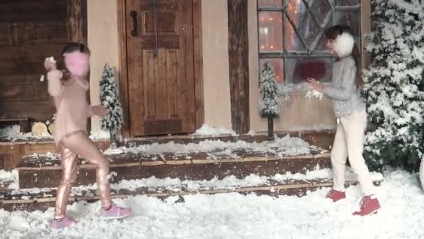 Χριστούγεννα ή Πρωτοχρονιά. τα παιδιά να διασκεδάσουν παίζοντας τεχνητό χιόνι. Χριστουγεννιάτικη διακόσμηση. Δύο μικρά κορίτσια στη βεράντα του σπιτιού. αργή κίνηση — Αρχείο Βίντεο