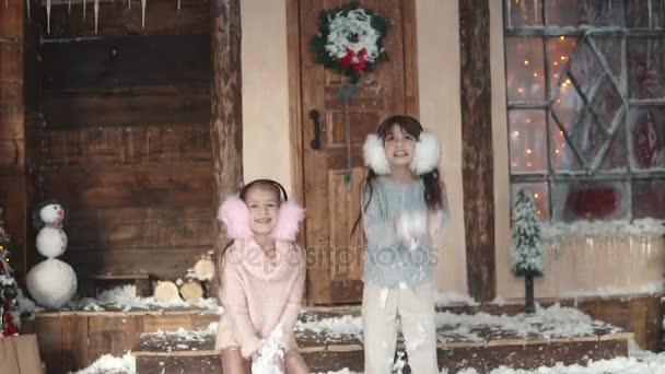 Noël ou Nouvel An. des enfants heureux vomissent de la neige. deux petites filles en casque de fourrure. Deux petites filles sur le porche de la maison. au ralenti — Video