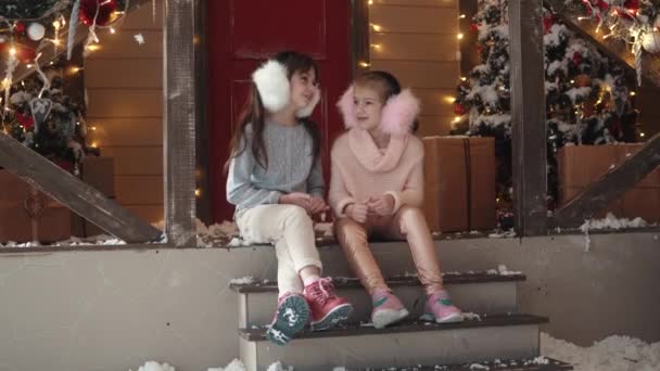 Neujahr oder Weihnachten. Kinder sitzen auf der Veranda vor dem Hintergrund des Neujahrsbaums und unterhalten sich — Stockvideo