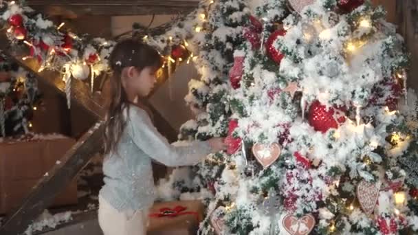 Χριστούγεννα ή Πρωτοχρονιά. παιδιά Κοιτάξτε γοητευτικά το δέντρο το νέο έτος. μικρό κορίτσι στα αυτιά γούνα κρεμάστε χριστουγεννιάτικα παιχνίδια για το χριστουγεννιάτικο δέντρο — Αρχείο Βίντεο