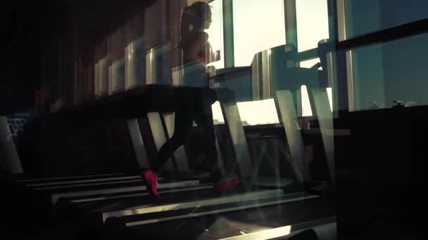 Cardio-training in de sportschool. silhouet van een meisje op een loopband. sportieve meisje draait op de loopband in de sportschool. atleet in sportkleding — Stockvideo