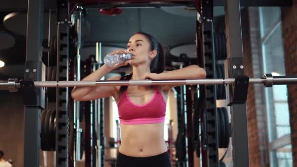 Chica sexy delgada es beber agua de una botella en el gimnasio. Deportiva en ropa deportiva descansando durante el entrenamiento — Vídeos de Stock