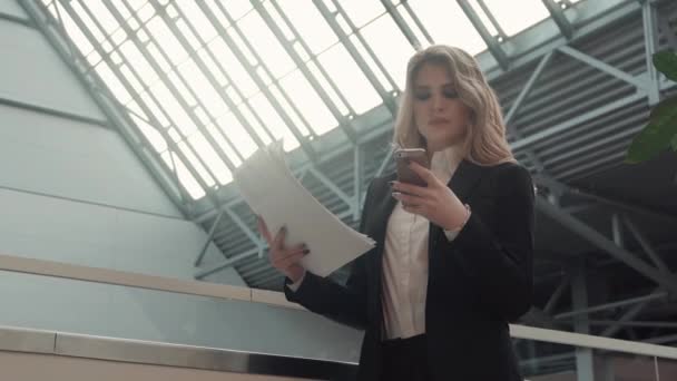 Mujer de negocios imprime un mensaje en un teléfono móvil mientras revisa los documentos. retrato de una mujer vestida de negocios en el vestíbulo de un edificio de oficinas — Vídeos de Stock