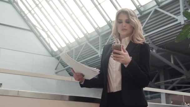 Üzletasszony egy üzenetet nyomtat egy mozgatható telefon dokumentumok ellenőrzése közben. portré egy nőt, az üzleti ruhákat a hallban egy irodaház — Stock videók