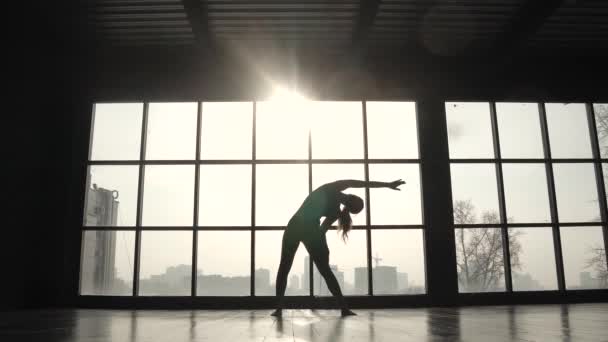 Silhuetten av en idrottsman mot bakgrund av ett stort fönster. Sport tjej gör uppvärmning före träning. ung kvinna praktisera yoga — Stockvideo