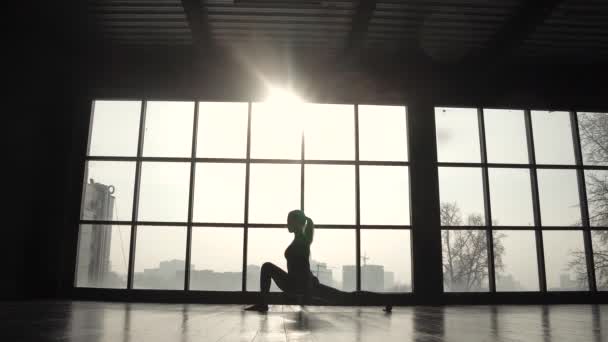 Silueta de un atleta sobre el fondo de una gran ventana. chica de deportes haciendo calentamiento antes del entrenamiento. mujer joven practicando yoga — Vídeo de stock