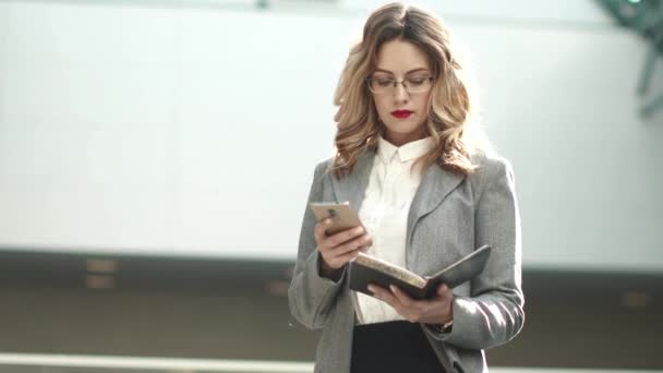 Mujer de negocios envía un mensaje desde el teléfono inteligente. la chica en un traje de negocios contesta el teléfono. retrato de una joven en el vestíbulo del centro de negocios — Vídeos de Stock