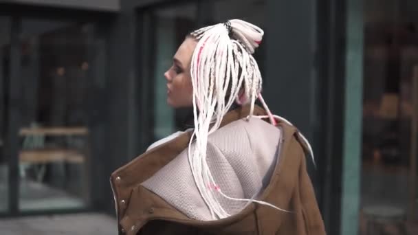 Chica con una apariencia inusual en el fondo de la arquitectura urbana. adolescente con rastas y piercing. una cultura juvenil moderna. Vista trasera. cámara lenta — Vídeos de Stock